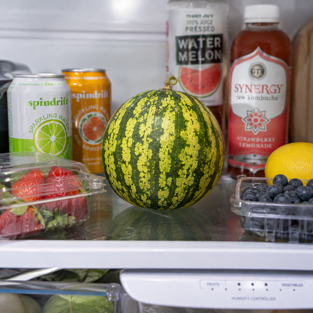 A small Sprouts and Petals Yellow Gem Sweet Watermelon rests on a refrigerator shelf, surrounded by strawberries, blueberries, a lemon, sparkling water cans, and bottles of watermelon juice and kombucha.